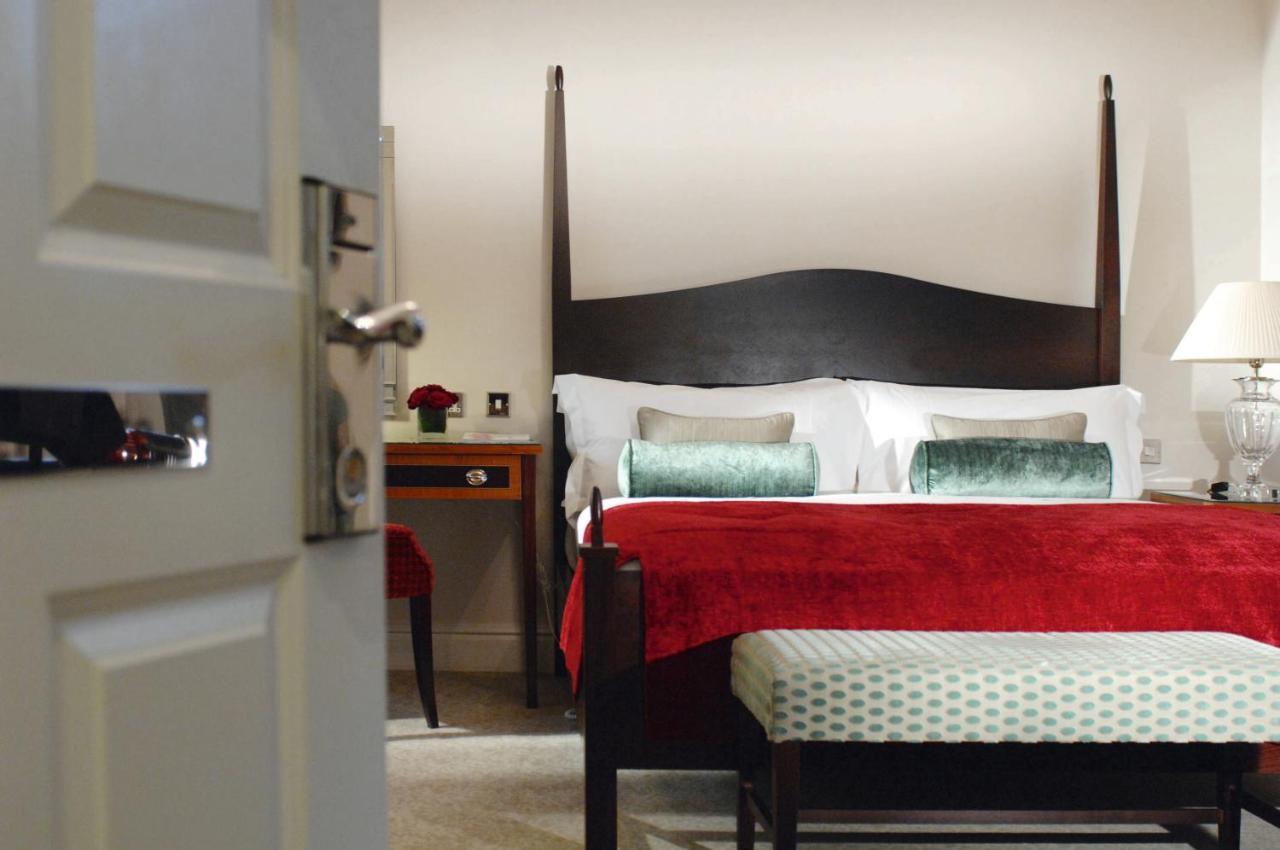 فندق ماكدونالد باث سبا الغرفة الصورة The photo shows a well-decorated hotel room. In the foreground, there is a partially opened door leading into the room. The focal point of the room is a large bed with a dark wooden headboard and stylish red and white bedding, accented with green pil