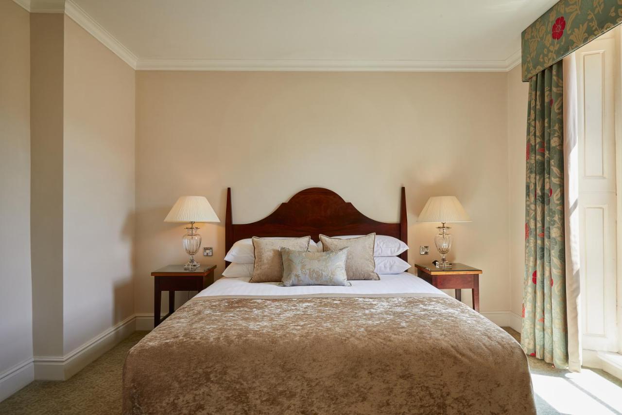 فندق ماكدونالد باث سبا المظهر الخارجي الصورة The photo shows a cozy hotel bedroom. It features a large bed with a plush, light-colored bedspread and several pillows. On either side of the bed, there are wooden nightstands with lamps providing soft lighting. The walls are painted a neutral color