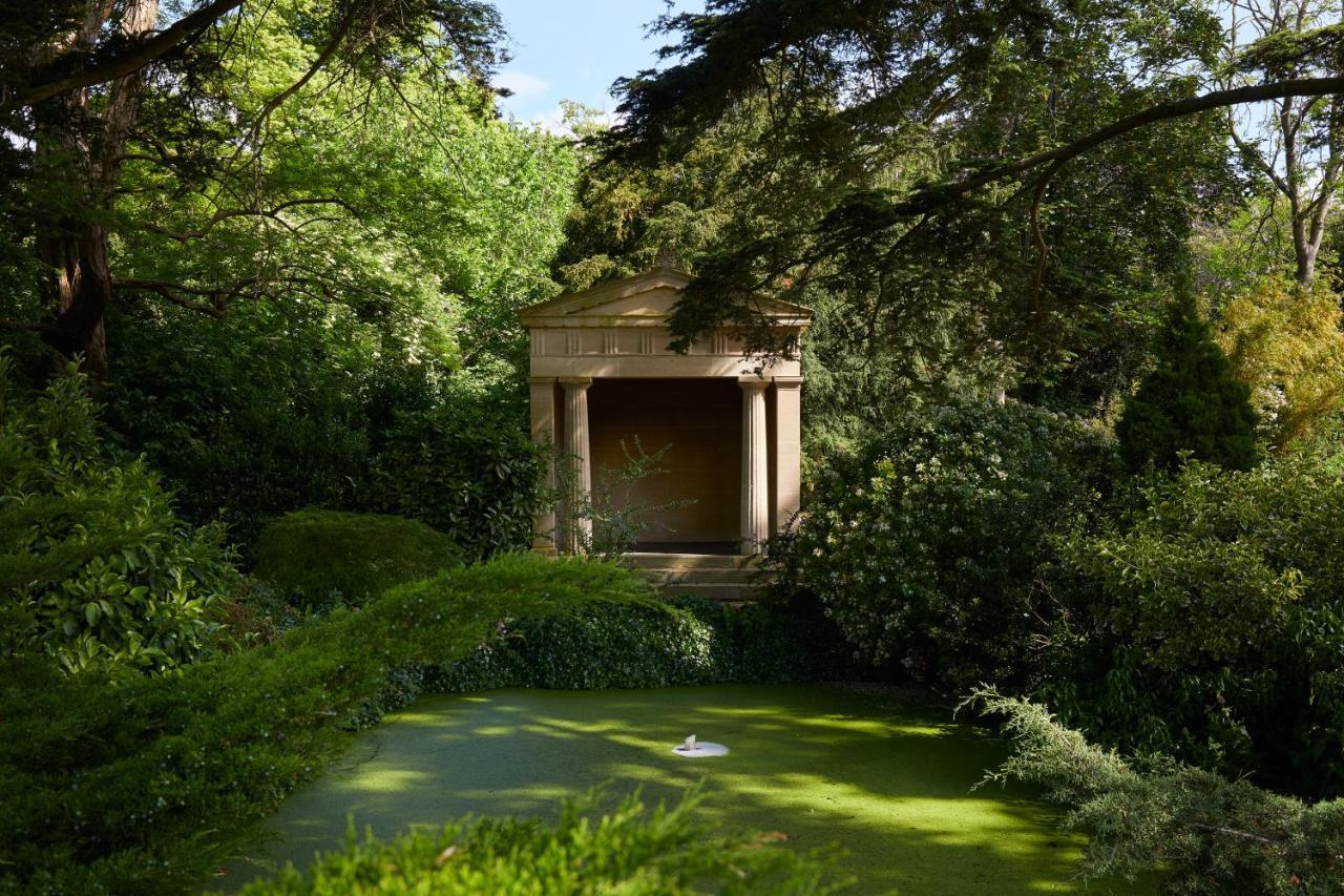 فندق ماكدونالد باث سبا المظهر الخارجي الصورة The photo shows a serene garden scene featuring lush greenery and a tranquil pond. In the background, there is a classical-looking structure with columns, resembling a small temple or pavilion. The pond appears to have vegetation covering its surface