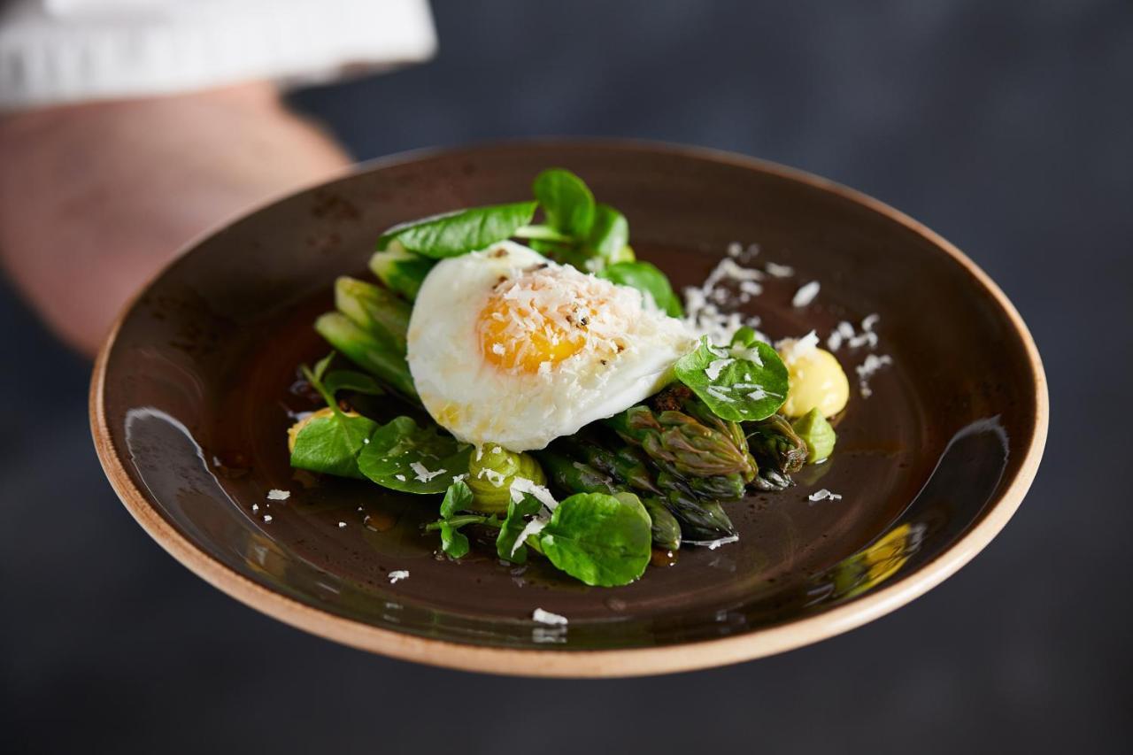 فندق ماكدونالد باث سبا المظهر الخارجي الصورة The photo shows a plate of beautifully arranged food, featuring a poached egg on top of a mix of fresh greens. The greens include asparagus, lettuce, and possibly some herbs, garnished with a sprinkle of grated cheese or a similar topping. The dish i
