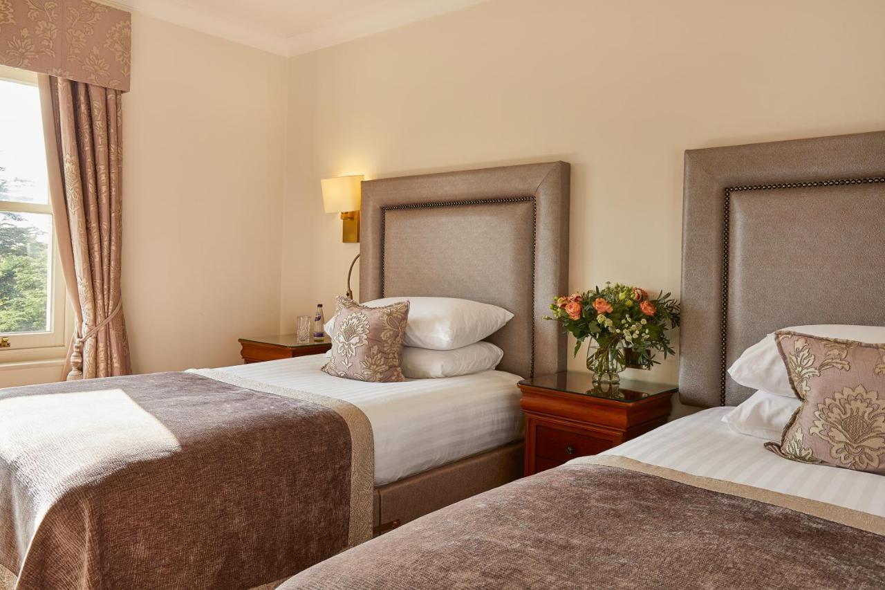 فندق ماكدونالد باث سبا المظهر الخارجي الصورة The photo shows a hotel room featuring two beds with elegant headboards. Each bed is neatly made with white linens and a soft, decorative throw blanket. There are matching pillows on the beds, and a small, stylish bouquet of flowers is placed on a wo
