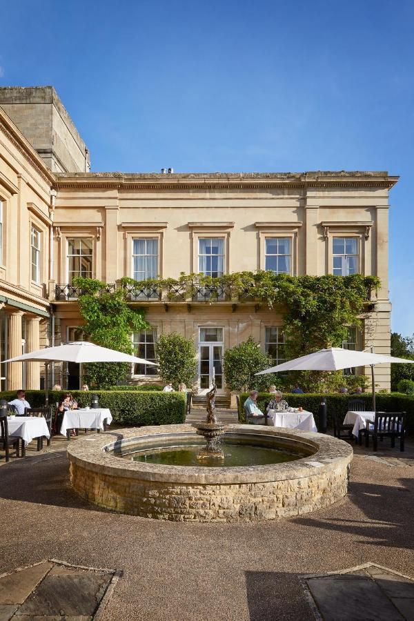 فندق ماكدونالد باث سبا المظهر الخارجي الصورة The photo depicts a charming outdoor dining area of a classic-looking building with elegant architecture. The structure features large windows and is adorned with greenery, including climbing plants. In the foreground, there is a circular fountain, a