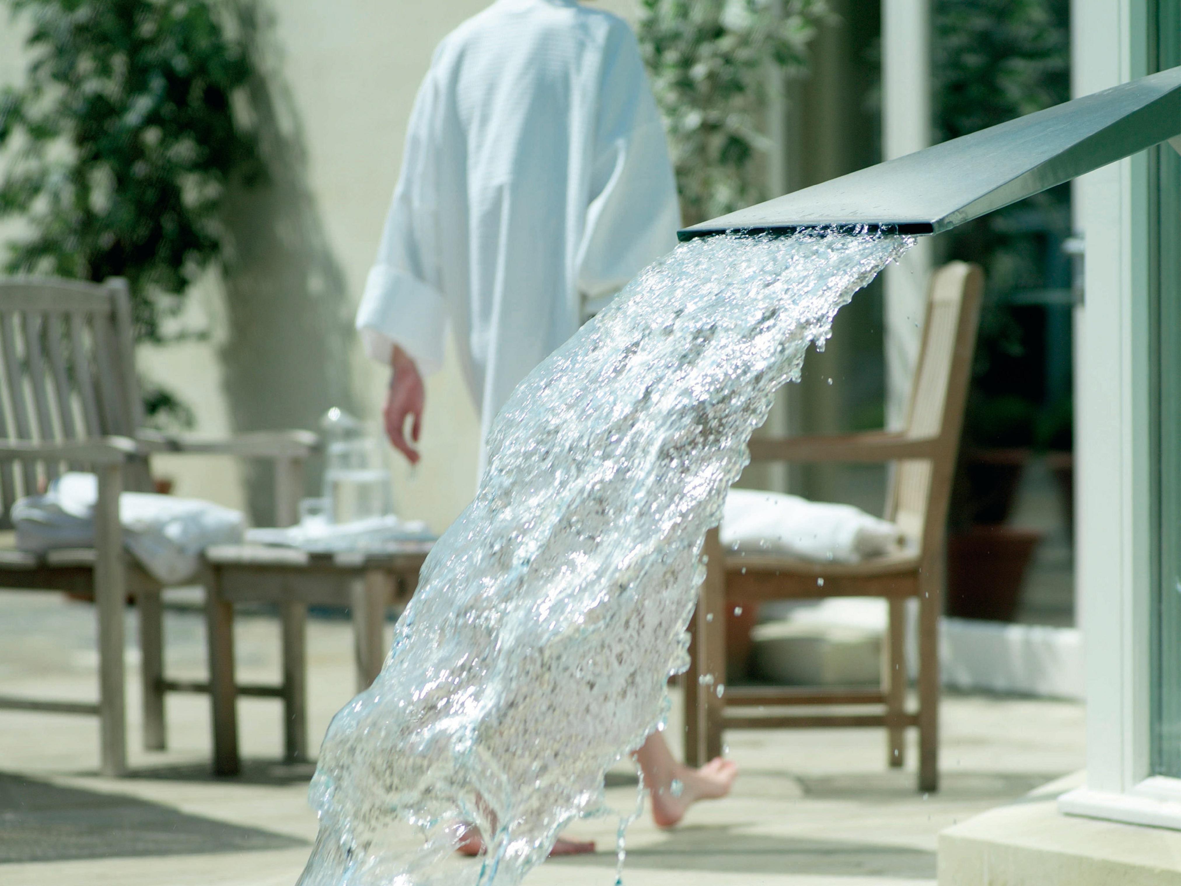 فندق ماكدونالد باث سبا المظهر الخارجي الصورة The photo shows a flowing water feature, with water elegantly cascading from a sleek spout. In the background, there is a person wearing a white bathrobe, walking away. Surrounding the area, there are wooden chairs and what appears to be a serene out