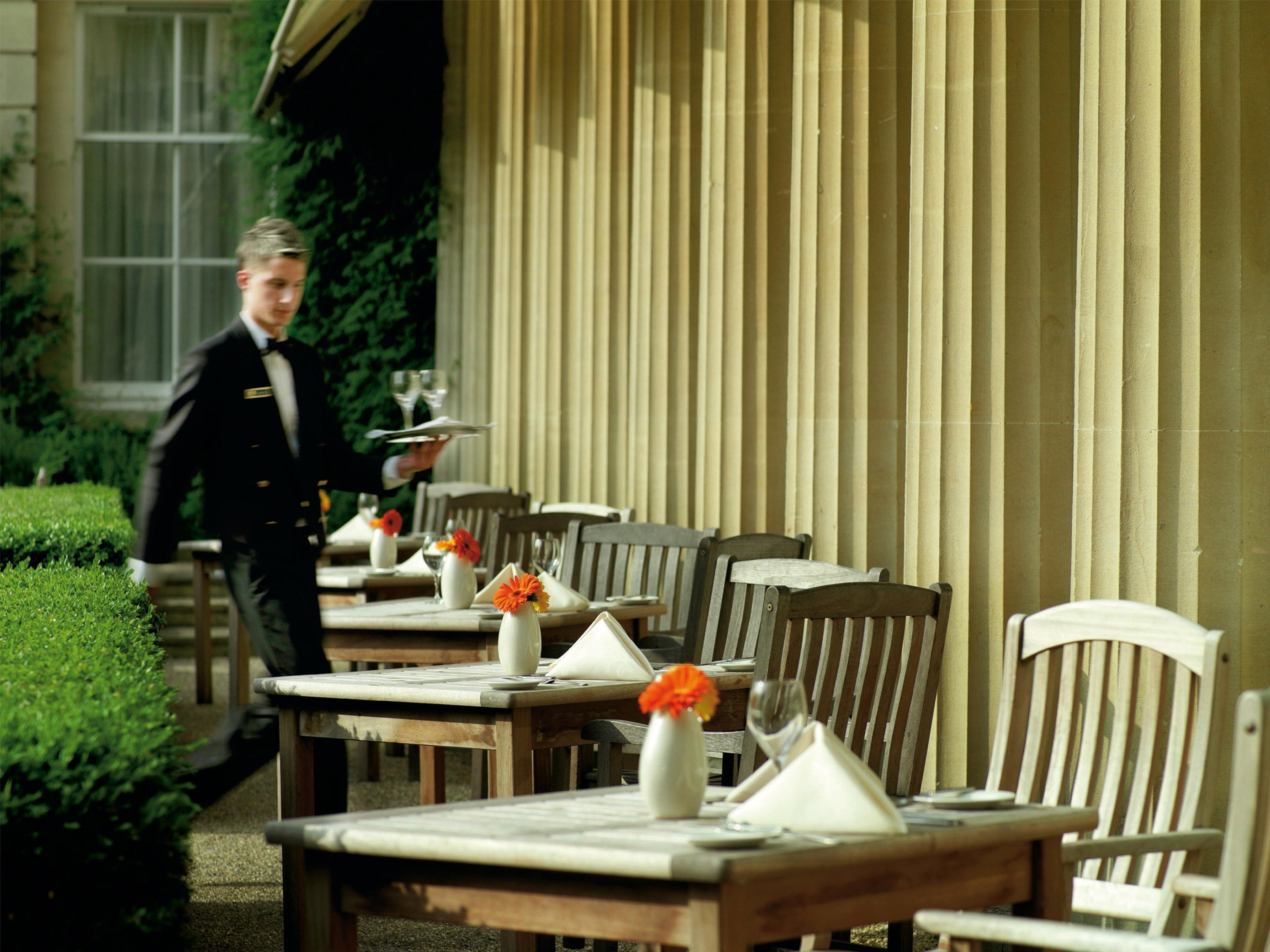 فندق ماكدونالد باث سبا المظهر الخارجي الصورة The photo shows an outdoor dining area, featuring several wooden tables and chairs arranged neatly. Each table is set with a white tablecloth and small vases containing flowers, adding a vibrant touch. A waiter in a formal black suit is seen moving t