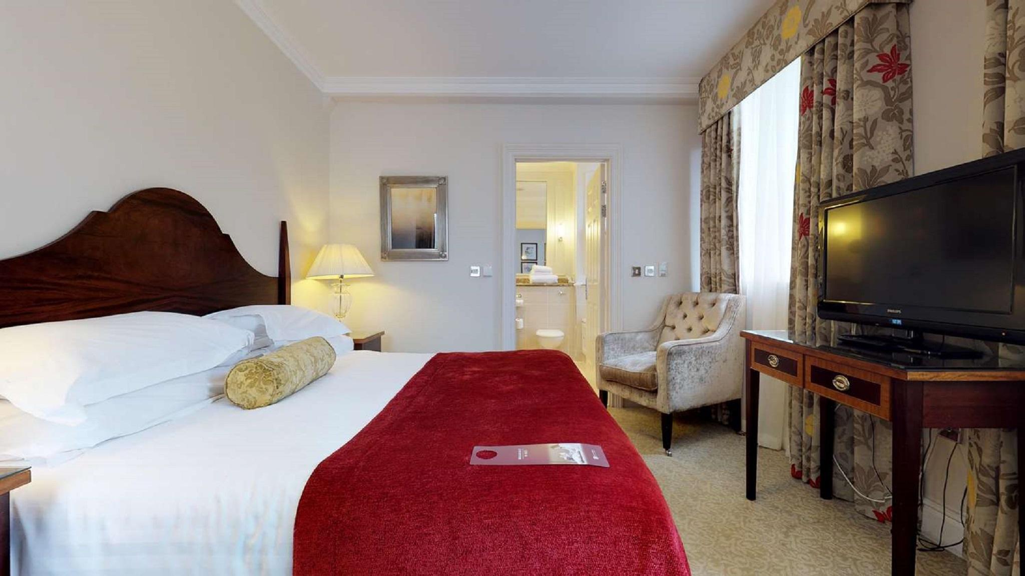 فندق ماكدونالد باث سبا المظهر الخارجي الصورة The photo shows a hotel room with a queen-sized bed covered in white linens and a deep red throw blanket. There is a decorative pillow on the bed. Next to the bed, there's a small nightstand with a lamp. In the background, a seating area features a b
