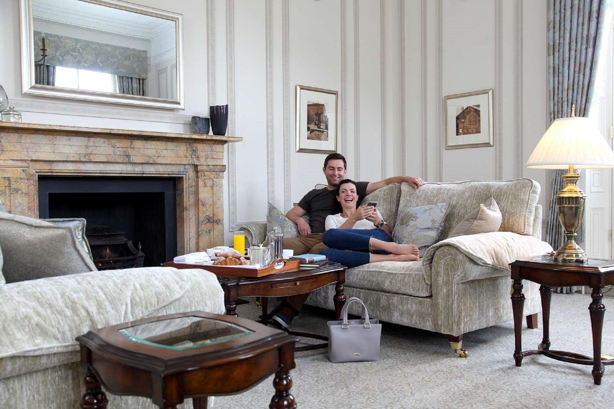 فندق ماكدونالد باث سبا المظهر الخارجي الصورة The photo shows a cozy living room setting where a couple is sitting together on a light-colored sofa. They appear relaxed, with the man sitting slightly behind the woman, who is smiling and looking at her phone. The room features elegant decor, incl