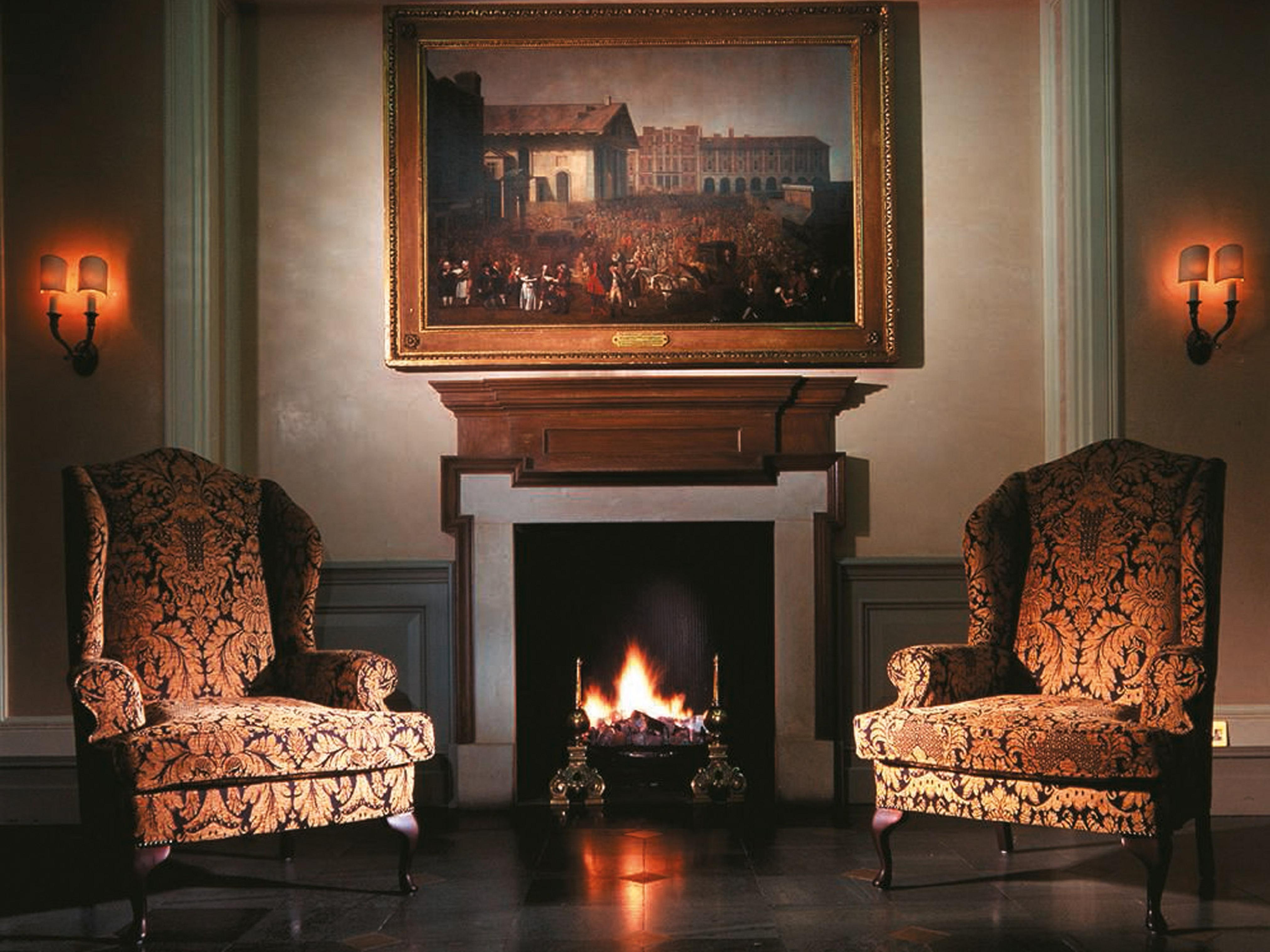 فندق ماكدونالد باث سبا المظهر الخارجي الصورة The image shows a cozy indoor setting featuring two ornate armchairs placed opposite a fireplace. Above the fireplace hangs a large painting in a decorative frame. The warm glow from the fire illuminates the room, and there are wall sconces providing