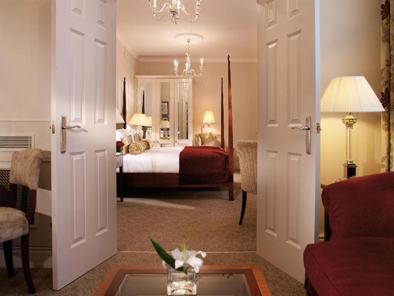 فندق ماكدونالد باث سبا الغرفة الصورة The photo shows a hotel room that can be viewed through open double doors. Inside, there is a neatly made bed with decorative pillows, a plush area rug, and soft lighting from lamps. The room features elegant furnishings, including a chair, a coffee 