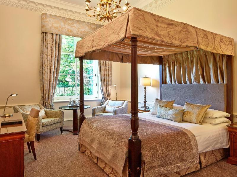 فندق ماكدونالد باث سبا الغرفة الصورة The photo shows an elegantly designed bedroom featuring a four-poster bed with a decorative canopy. The bed is dressed with luxurious bedding and pillows, creating a cozy and inviting atmosphere. Flanking the bed are two comfortable armchairs and a s