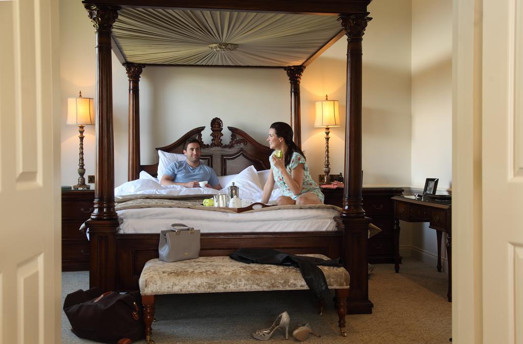 فندق ماكدونالد باث سبا المظهر الخارجي الصورة The image depicts a spacious, well-lit bedroom featuring a large four-poster bed with an ornate headboard. A man and a woman are sitting on the bed, appearing to engage in conversation. The woman is dressed in a patterned outfit, while the man is wea